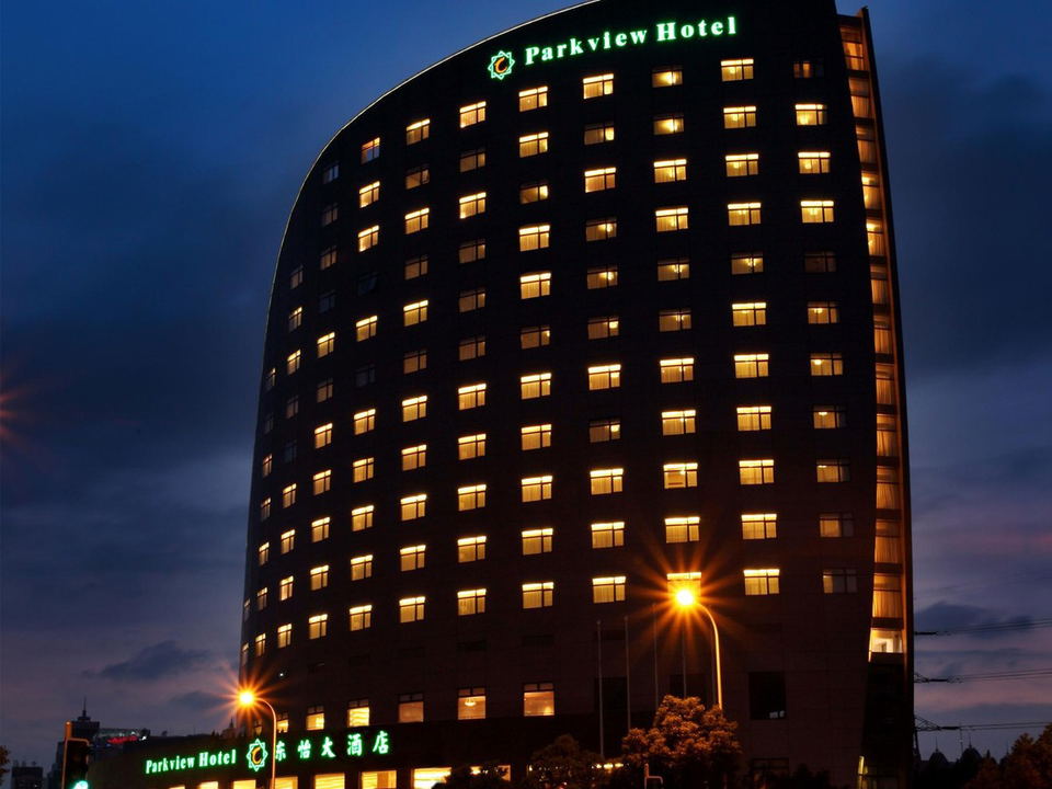 A nighttime exterior view of the Parkview Hotel in Shanghai, with its illuminated name in green on the curved facade and light trails from passing