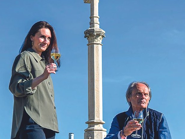 Hombeline Guyon and a companion with wine glasses and a bottle, near a stone monument.