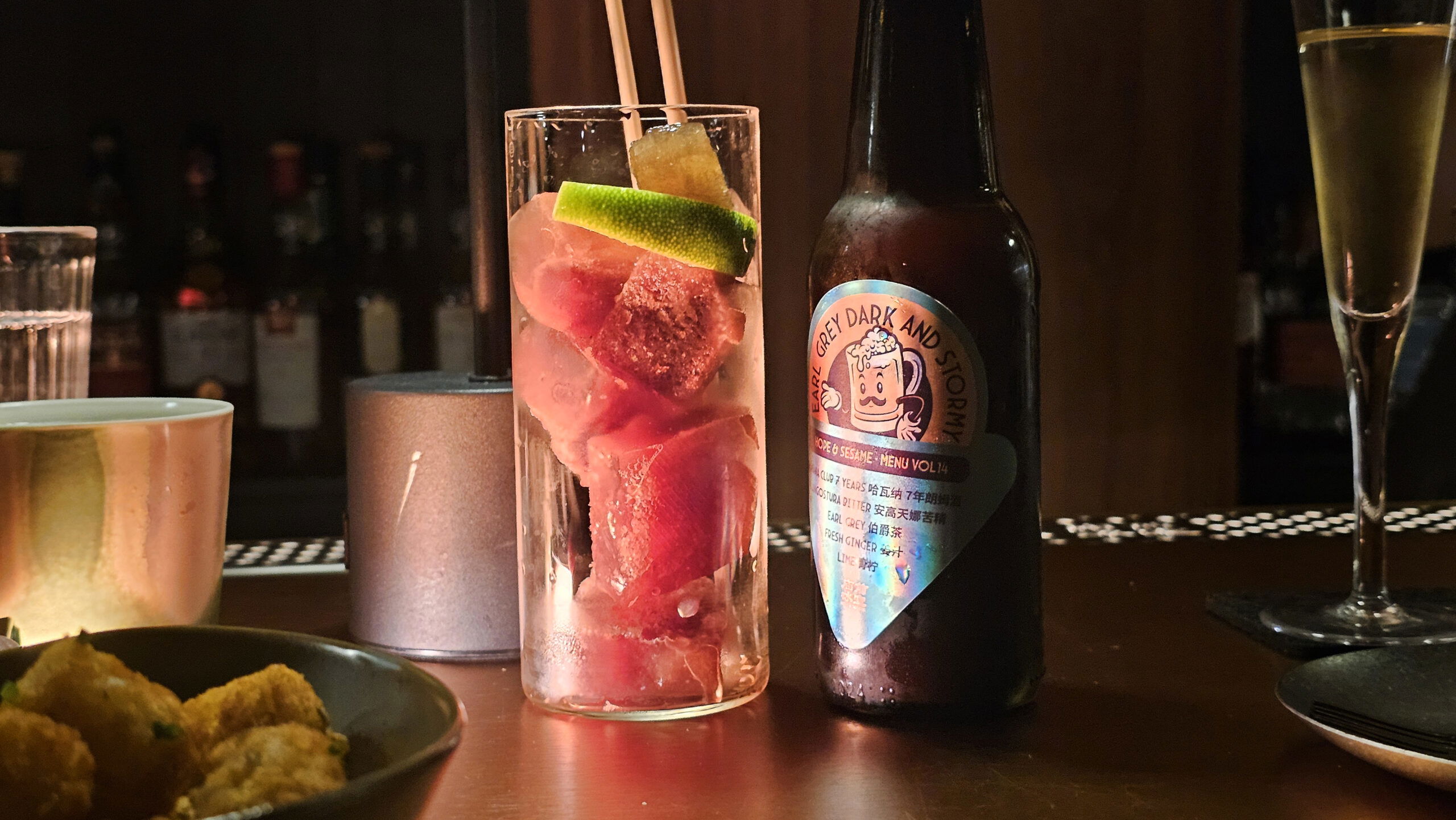 A tall glass with red ice cubes and a lime wedge, next to a bottle labeled "Grey Dark and Stormy" on a bar counter.