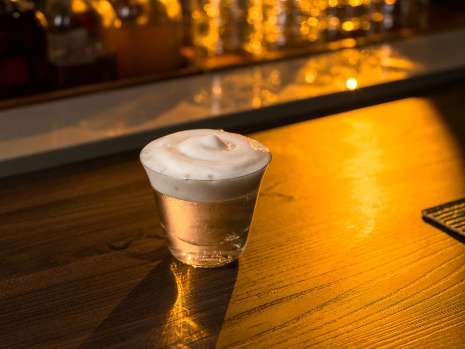 A foamy cocktail on a wooden bar, reflecting the warm glow of the lights.