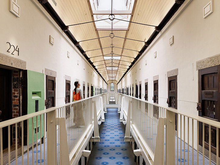 A long, symmetrical corridor with white railings, dark cell doors, exposed beams, skylights, and patterned blue tile flooring.