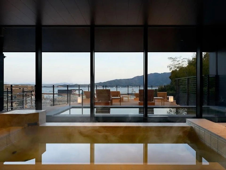 An indoor onsen at Hotel Fork & Knife Miyajima, with floor-to-ceiling glass walls opening to an outdoor terrace with wooden lounge chairs and a view