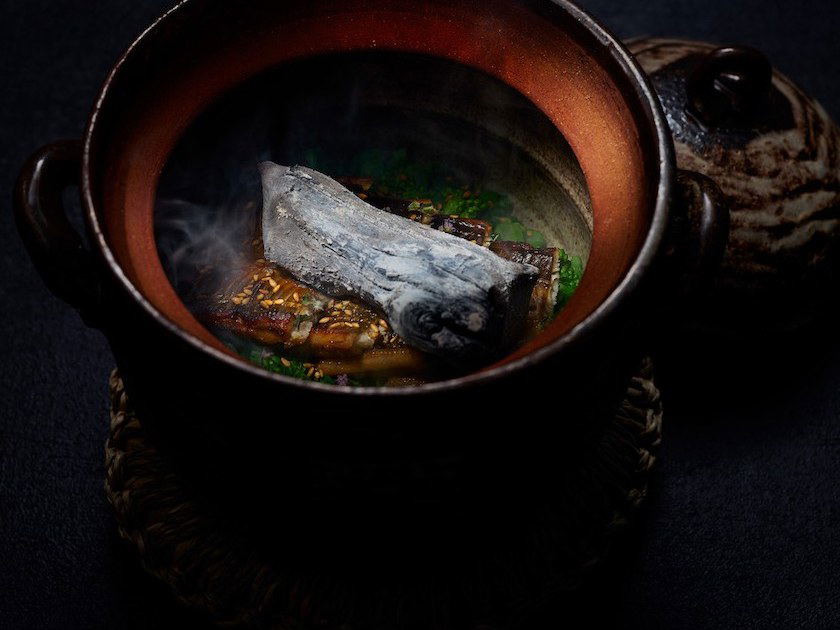 A dark, dramatically lit earthenware pot containing conger eel with steam rising, garnished with green herbs and sesame seeds.