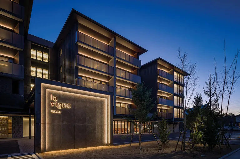 A contemporary multi-story hotel building with dark exterior and glowing windows photographed at twilight, with bare trees visible to the right.