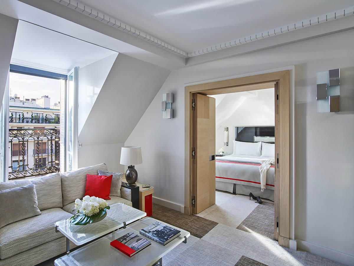 A bright Art Deco hotel suite living room with a beige sofa, glass coffee table, and an open doorway leading to a bedroom.