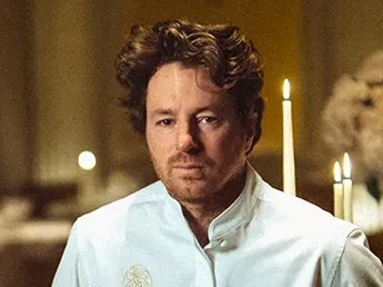 Chef Jean Imbert in a white chef's jacket stands in an elegant, dimly lit dining room with candles, flowers, and formal table settings.