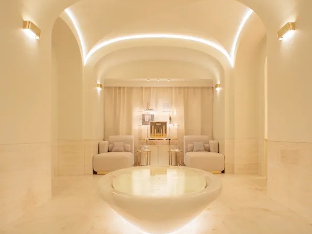 A luxurious spa interior with vaulted ceilings, integrated LED lighting, cream-toned marble, symmetrical seating, and a central circular feature.