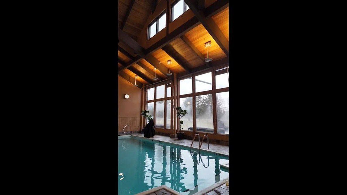 An indoor swimming pool with a high, vaulted wooden ceiling, large windows, and warm pendant lighting.