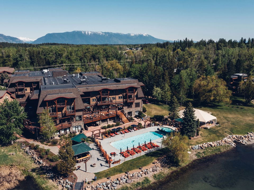 An aerial view of Hotel Whitefish, a large timber-frame lodge with an outdoor pool, hot tub, and bar, on a lakefront with mountains.