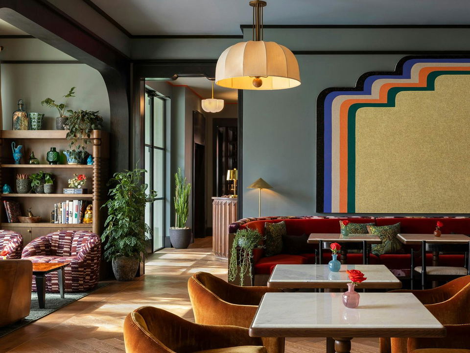 A stylish hotel bar interior with dark sage-green walls, walnut shelving, red velvet banquette seating, amber velvet chairs, and marble-top tables.