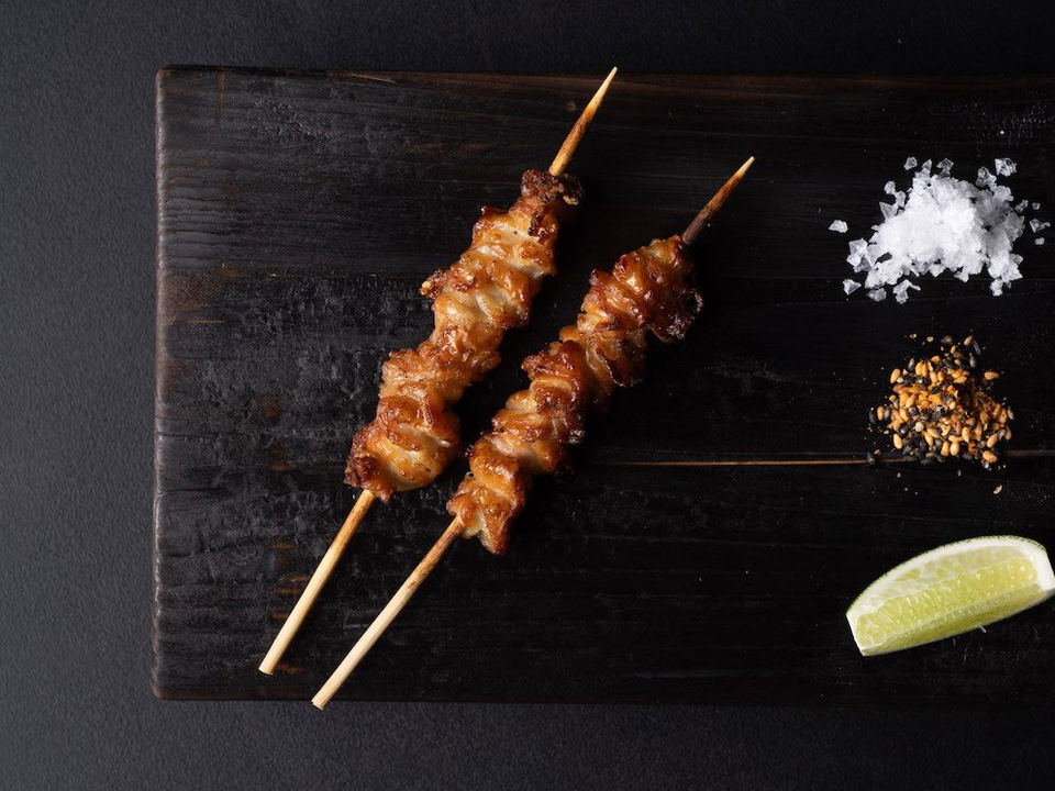 Two grilled yakitori skewers on a dark wooden board with flaky salt, sesame seeds, and a lime wedge, presented by Comma Hospitality.