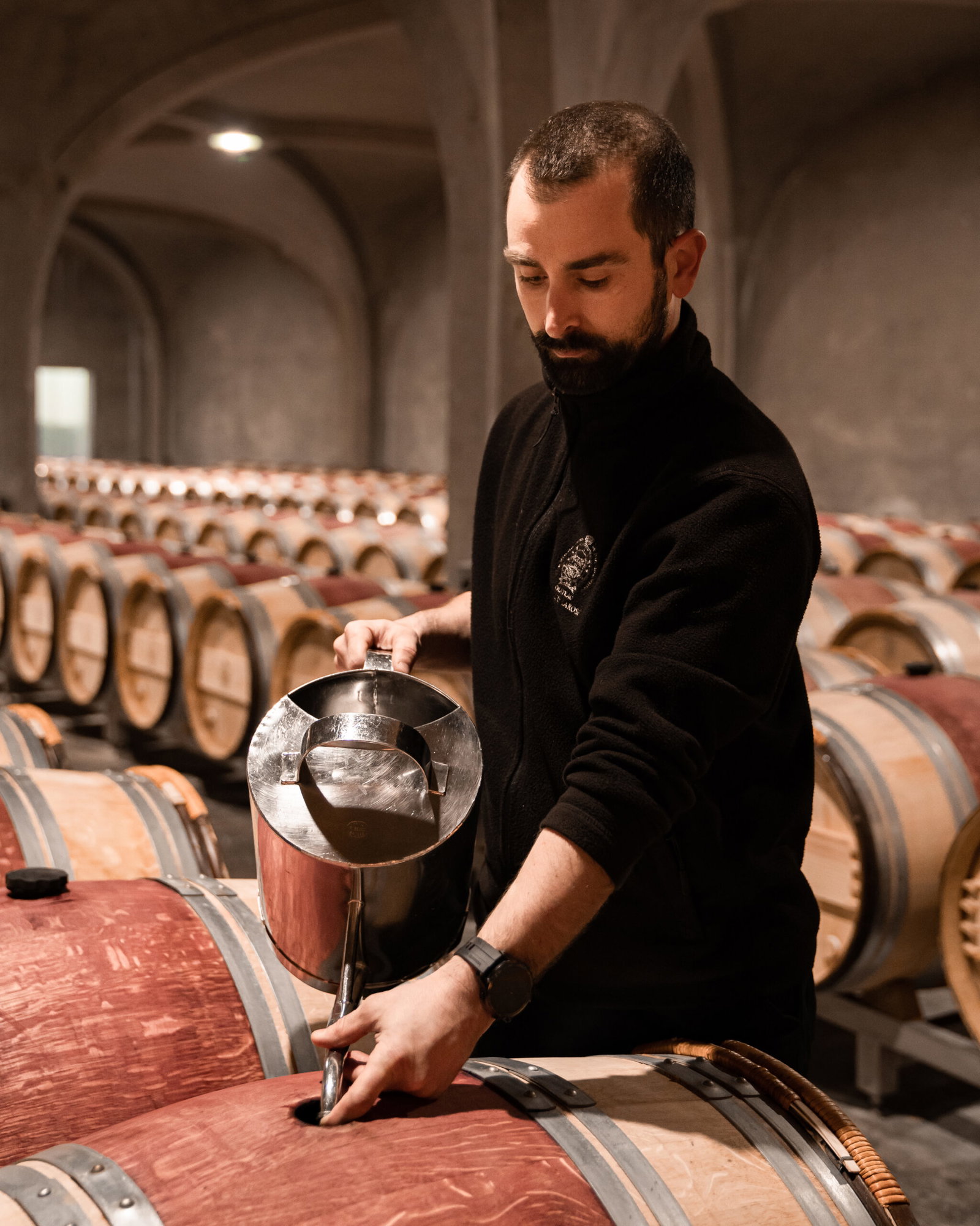 A man in black attire drawing a wine sample from an oak barrel in a vaulted cellar with rows of barrels