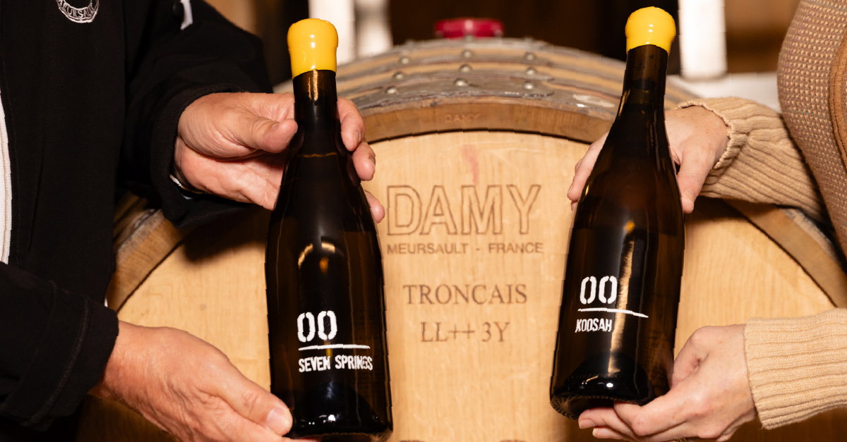 Two people holding dark wine bottles with yellow caps in front of a wooden barrel marked 'DAMY MEURSAULT - FRANCE TRONCAIS LL++3Y'