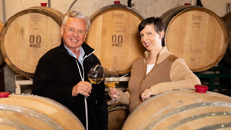 Two people toasting with white wine glasses in a barrel cellar with visible '00 WINES' branding on barrels behind them