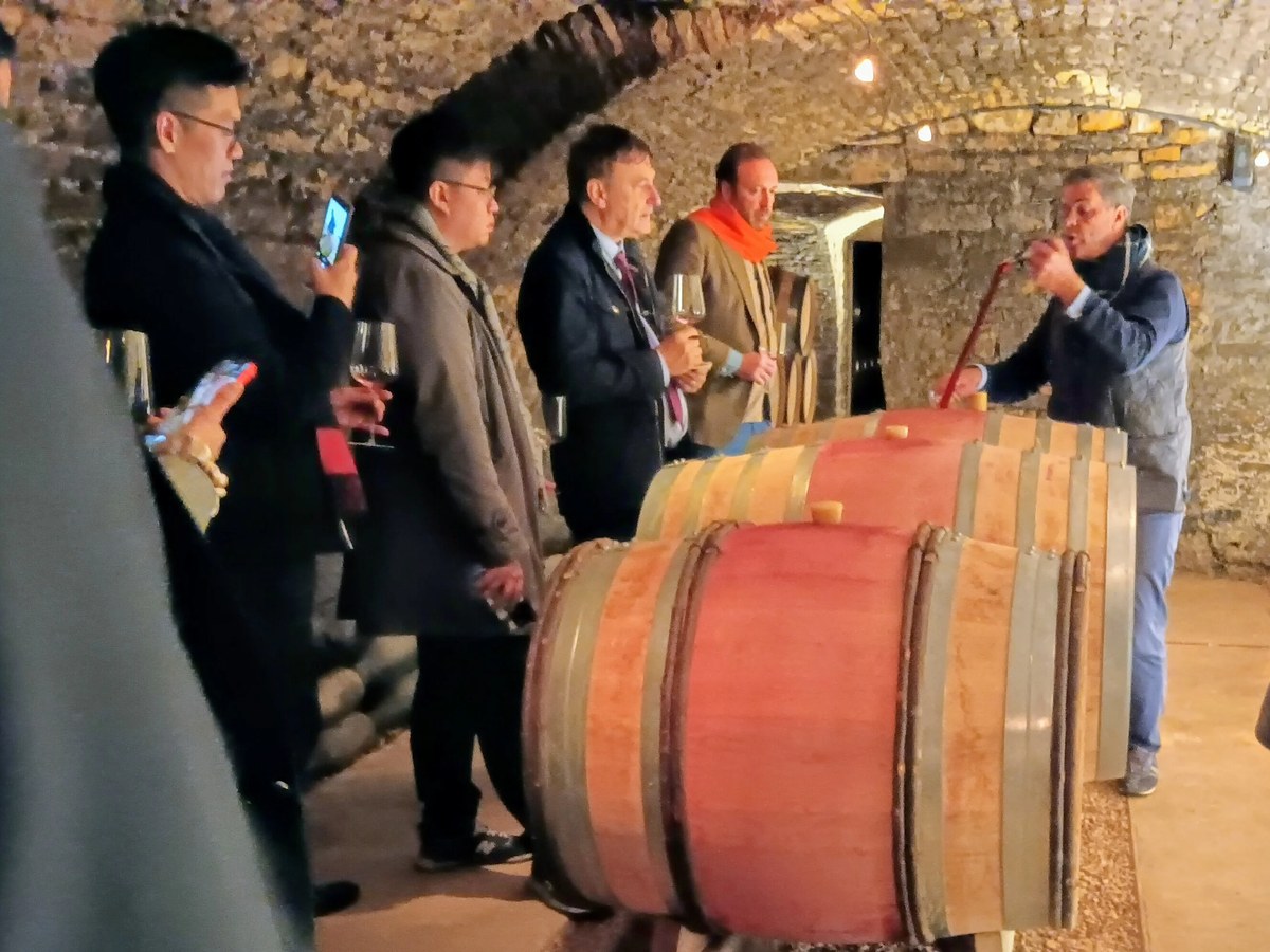 A group of people in a low-lit, stone-vaulted cellar, gathered around wooden wine barrels, with one man using a wine thief.