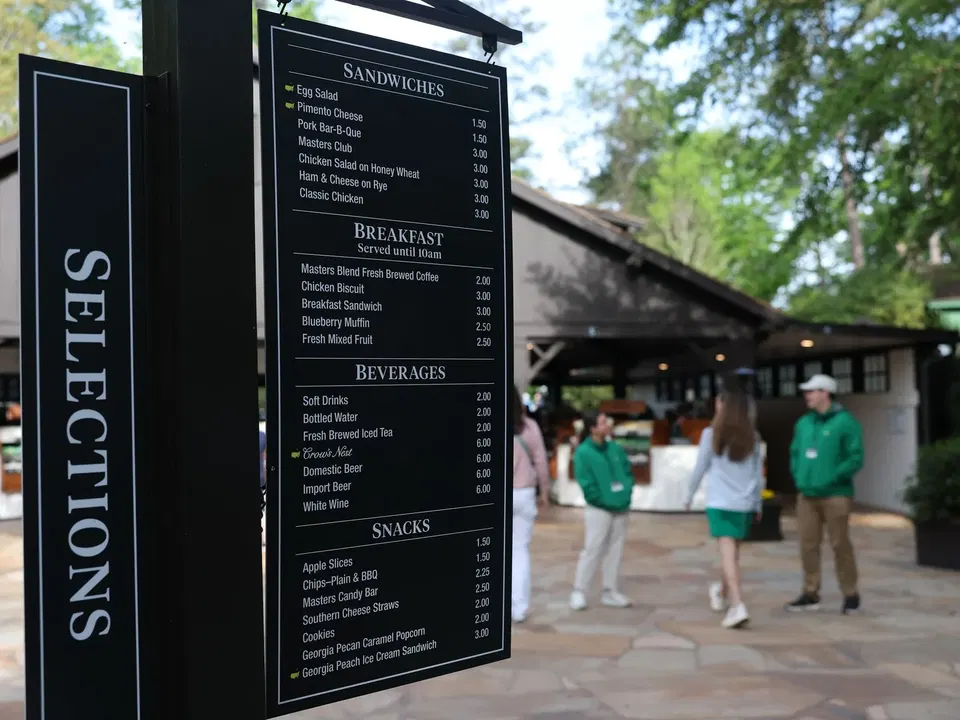 A black menu board showing sandwiches, breakfast items, beverages, and snacks with prices, with spectators and buildings visible in the background.