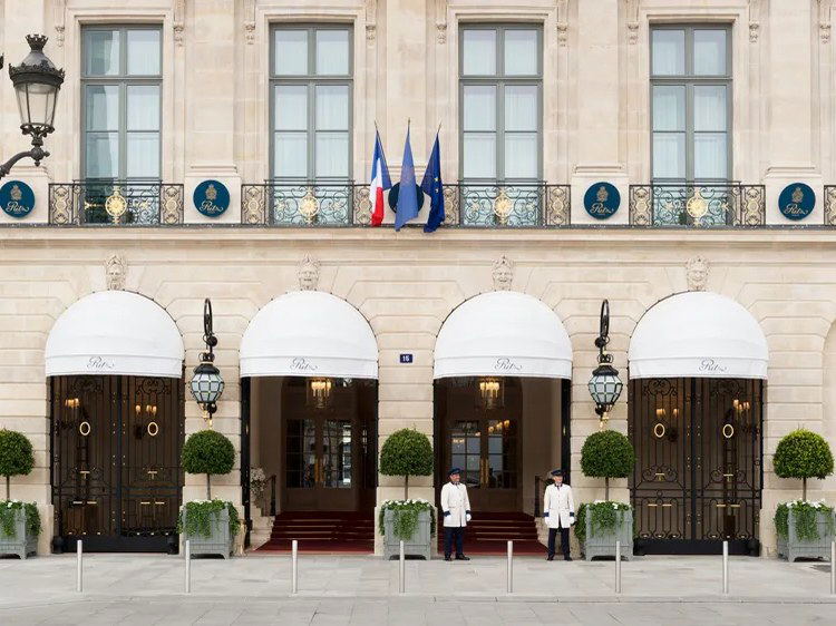 The exterior of the Ritz Paris hotel, featuring white dome awnings over entrances, teal Ritz medallions on a balcony, and two doormen.