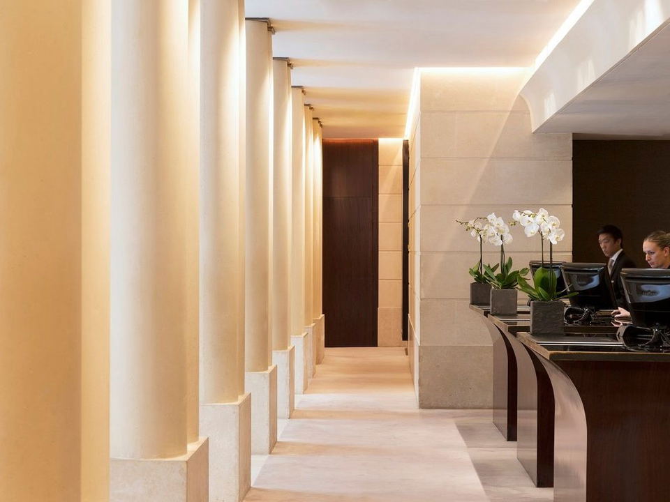 Two hotel staff members, one man and one woman, work at a dark wood reception desk with computers and white orchid plants.