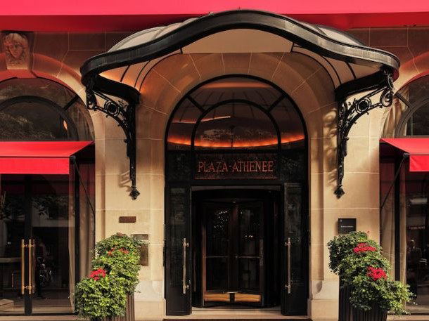 The exterior of Hotel Plaza Athenee Paris, featuring its main entrance with an arched awning, ornate ironwork, and red awnings.