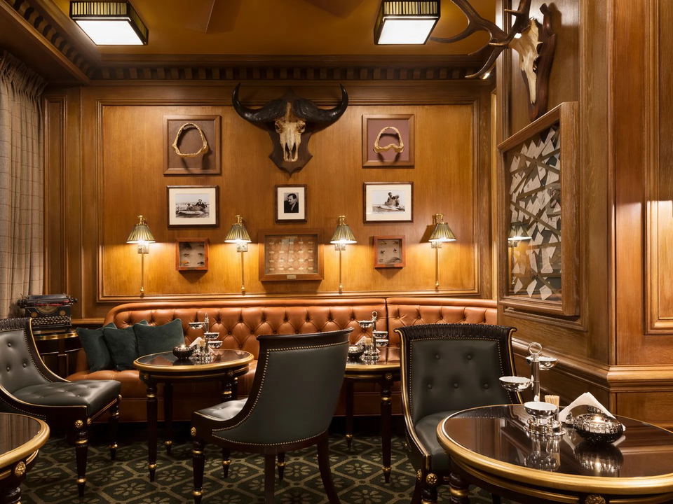 An intimate, wood-paneled bar lounge interior with tufted leather banquettes, brass sconce lighting, and animal trophy mounts.