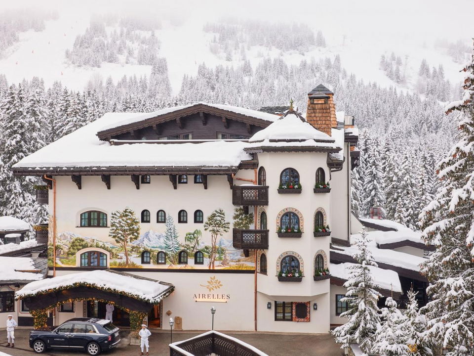 An aerial view of Hotel Les Airelles Courchevel, a white Alpine chalet with a painted mural, snow-covered roof, and 'AIRELLES' signage, surrounded.