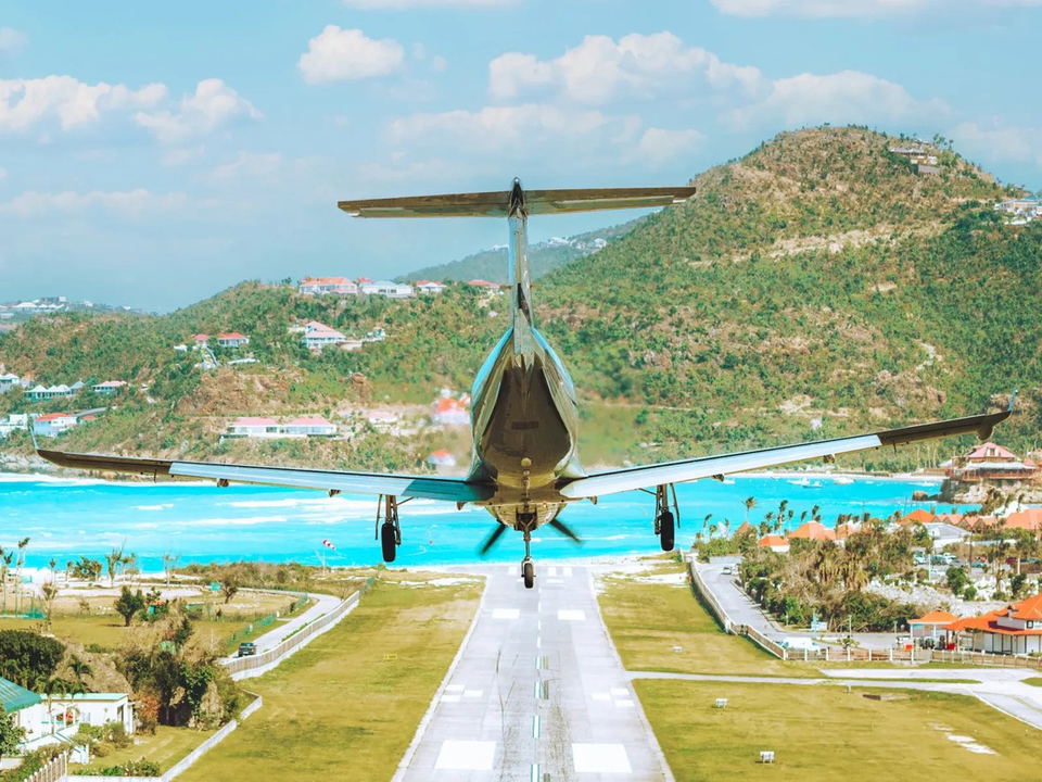 Small aircraft landing on a runway with turquoise water and hills in background.
