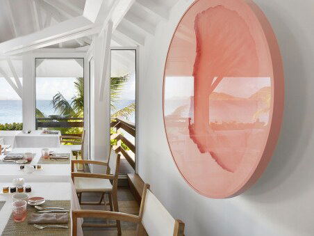 A white dining room with wooden chairs, a pink circular wall art piece, and large windows overlooking palm trees and ocean views.