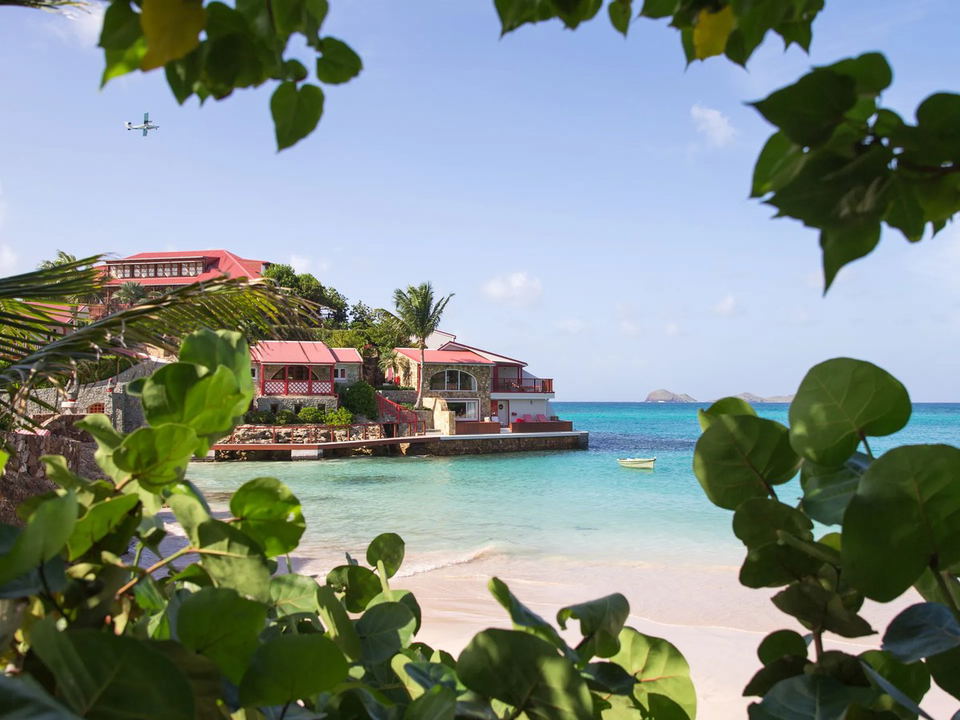 A small plane flies over the red-roofed buildings of Eden Rock St. Barths, perched on a rocky promontory above a turquoise bay with a white sand.