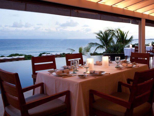 An open-air dining terrace at dusk, featuring a long table set with white linens, candles, and dark wooden chairs, overlooking a calm ocean.