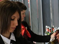 A woman in professional attire holds and examines a wine glass while tasting wine.