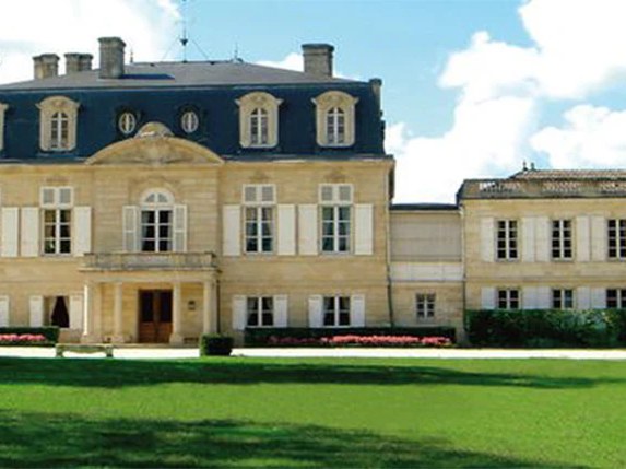 A grand, light-colored stone château with a dark mansard roof and white-shuttered windows, surrounded by green lawns and trees under a blue sky.