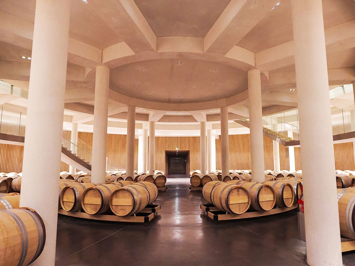 A grand, circular barrel cellar with tall white columns, a vaulted ceiling, and rows of oak wine barrels on low platforms, with wood-slat walls and
