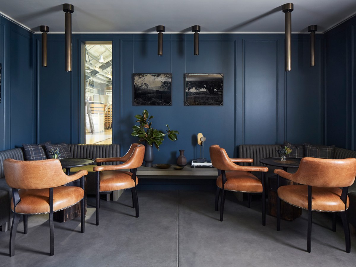 A sophisticated tasting room interior with deep blue paneled walls, tan leather armchairs, and a glimpse into a barrel hall.
