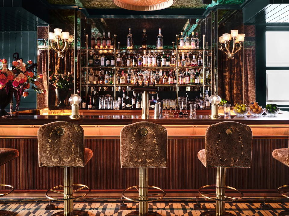An opulent, dimly lit cocktail bar interior featuring ornate velvet barstools with gold bases, a warm-lit back bar stocked with hundreds of bottles,