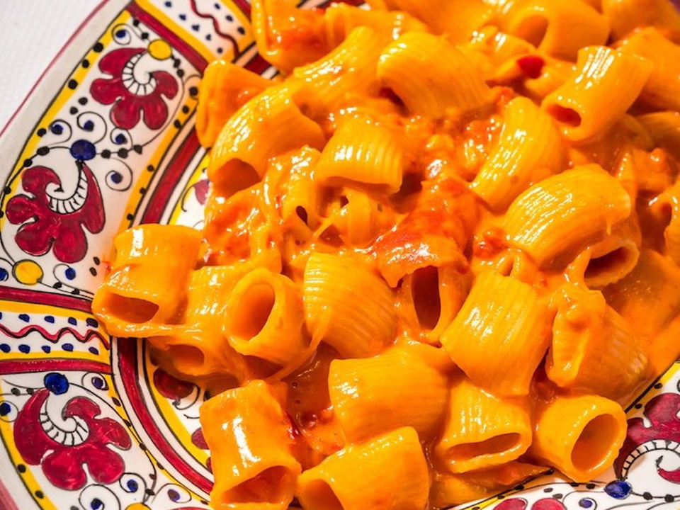 A close-up of rigatoni pasta coated in a vibrant orange-red vodka sauce, served in a hand-painted ceramic bowl with red, yellow, and blue patterns.