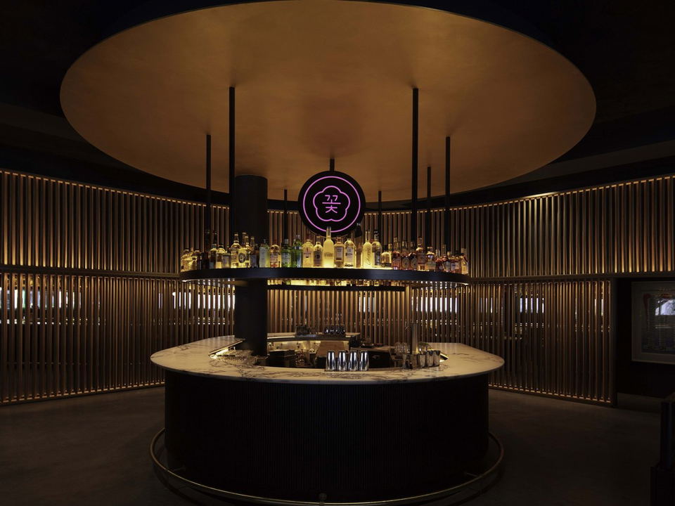A circular bar with a marble top and a black fluted base, surrounded by a golden slatted wall, under a large circular golden ceiling canopy.