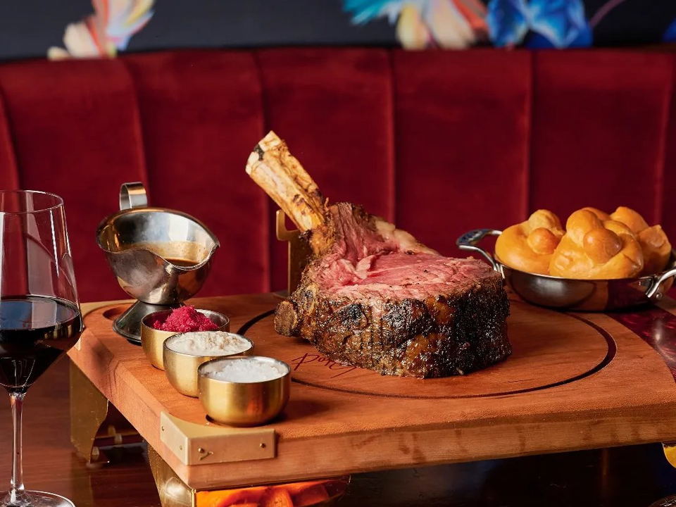 A large bone-in prime rib roast on a branded wooden carving board with gold condiment cups, a gravy boat, dinner rolls, side dishes, and a glass of