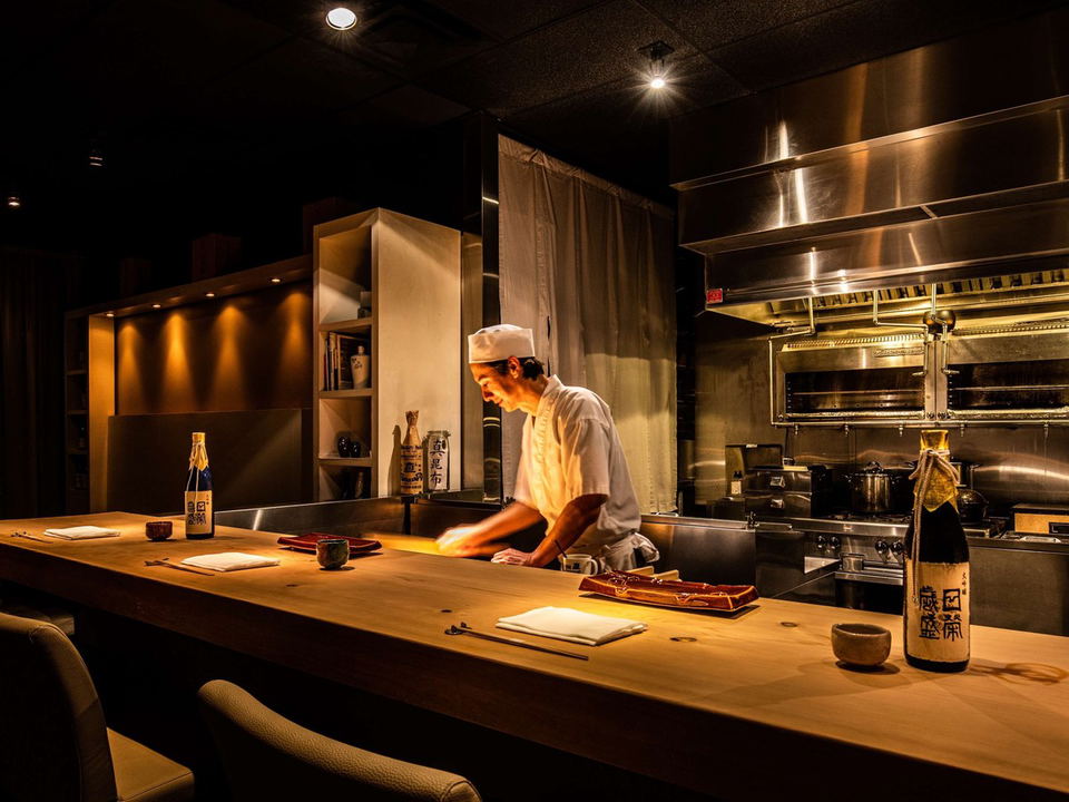 A chef in a white uniform prepares food behind a long, light wooden counter in a dimly lit, intimate restaurant.