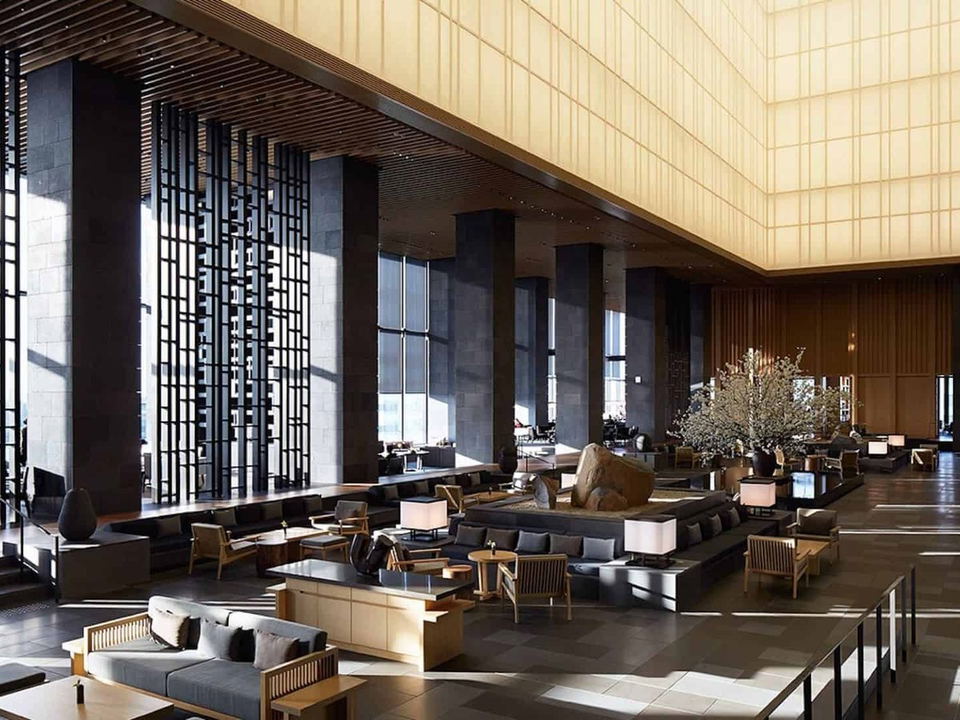A grand, double-height hotel lobby with dark stone columns, wooden lattice screens, and warm backlit ceiling panels, featuring low seating.