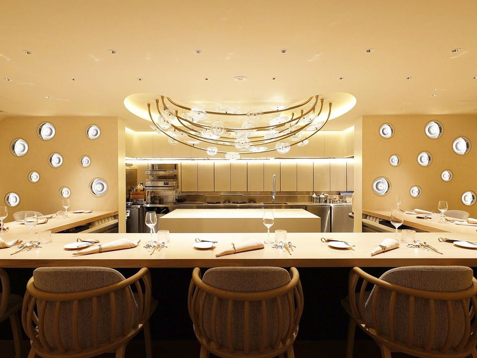 An elegant, warm-toned open-kitchen counter dining room with a distinctive crescent-shaped chandelier, circular wall-mounted decorative plates, and a
