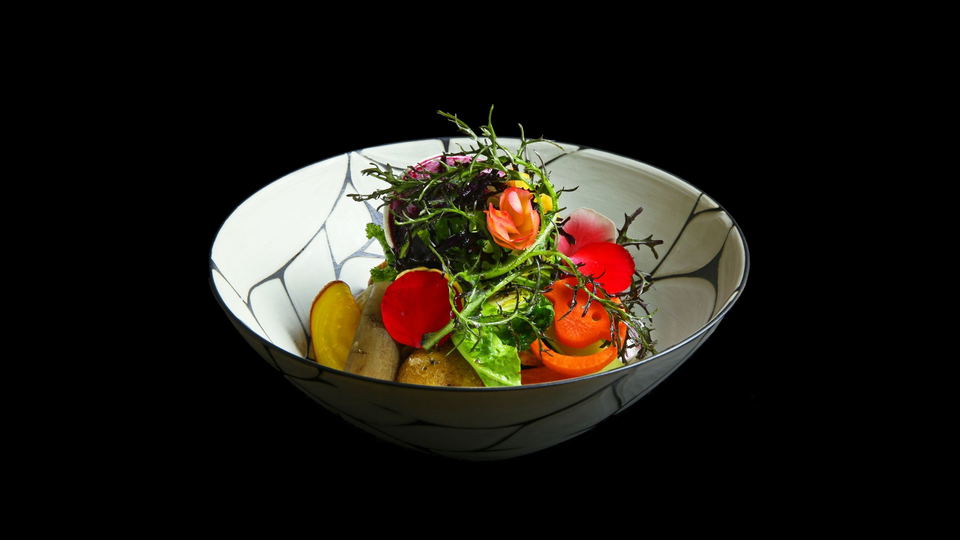 A white ceramic bowl with black geometric line patterns contains a composed dish with red and orange tomatoes, dark leafy greens, delicate microgreens, and what appears to be a protein element on a black background.