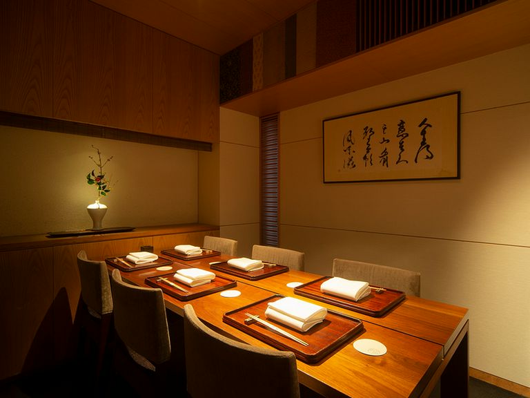 A private dining room with a wooden table set for six guests, featuring traditional Japanese kaiseki place settings on lacquered trays, warm wood pane