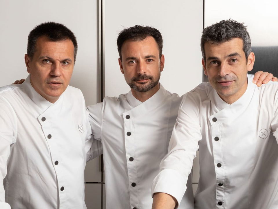 Three men wearing white chef's coats with black buttons stand side by side against a white wall with metal fixtures.
