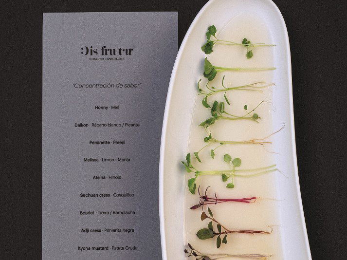 A white platter holds a row of microgreens and herb sprouts, next to a grey Disfrutar Restaurant menu card titled "Concentración de sabor."
