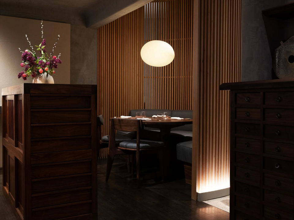 A dimly lit, intimate dining room at n/naka, featuring warm wood slatted walls, a round pendant light, and a set dining table.
