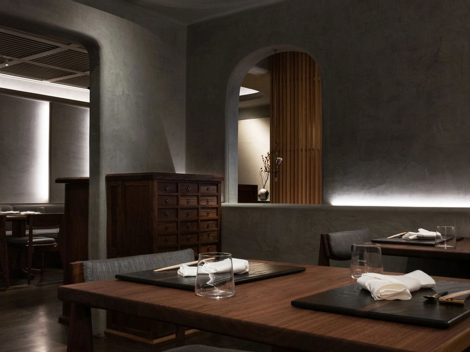 Dimly lit, elegantly appointed dining room with dark wood tables set with black placemats, chopsticks, folded white napkins, and clear glassware.