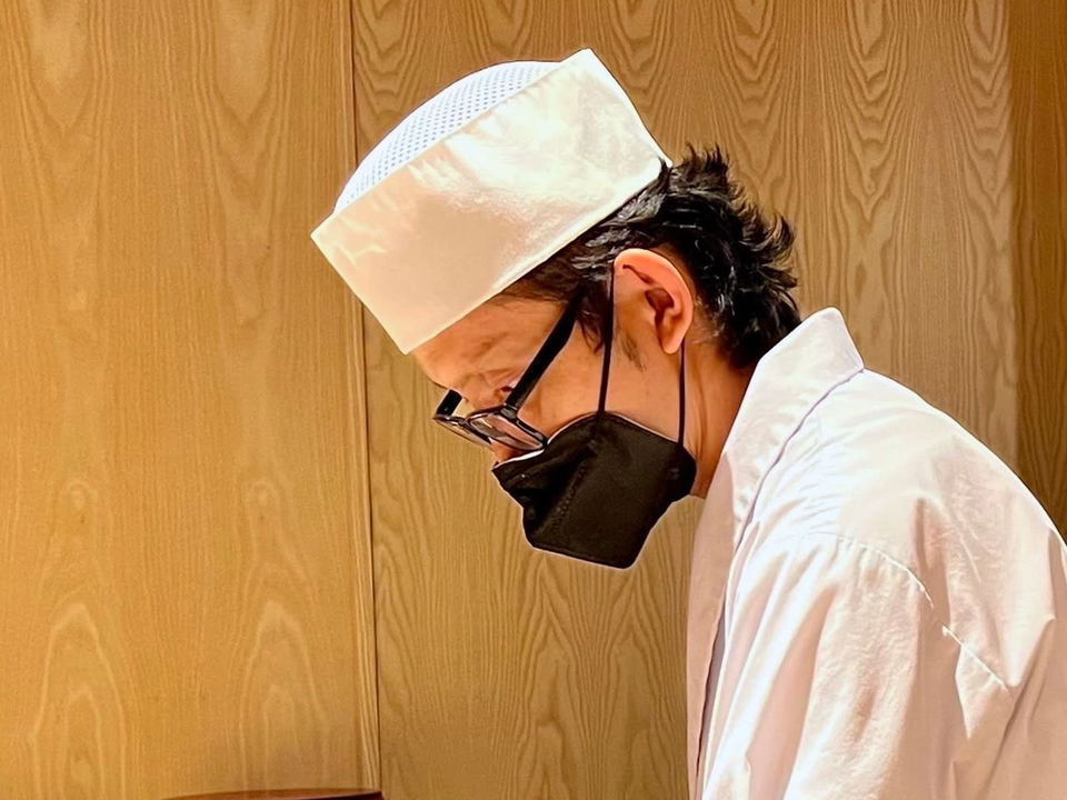A Japanese chef in a white uniform and black mask slices fish at a wooden omakase counter, with various cuts of fresh fish displayed in
