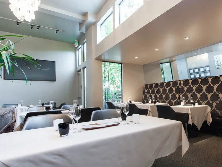 A modern, upscale restaurant dining room with white-tablecloth tables, wine glasses, dark patterned banquette seating, and high windows.