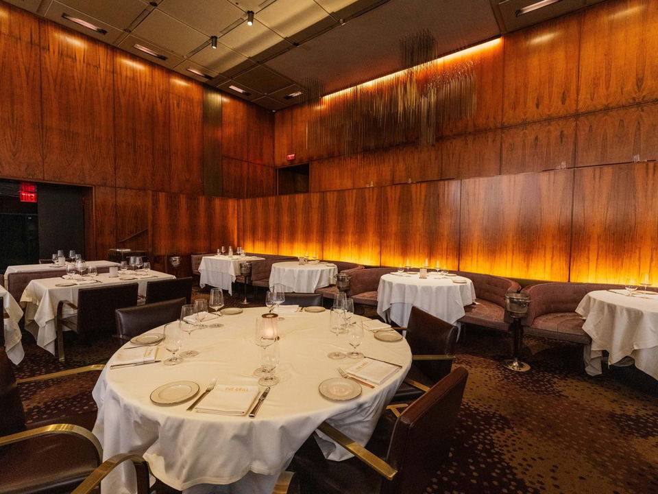 The interior of The Grill, an elegant dining room with wood-paneled walls, white-tableclothed round tables, and plush banquette seating.