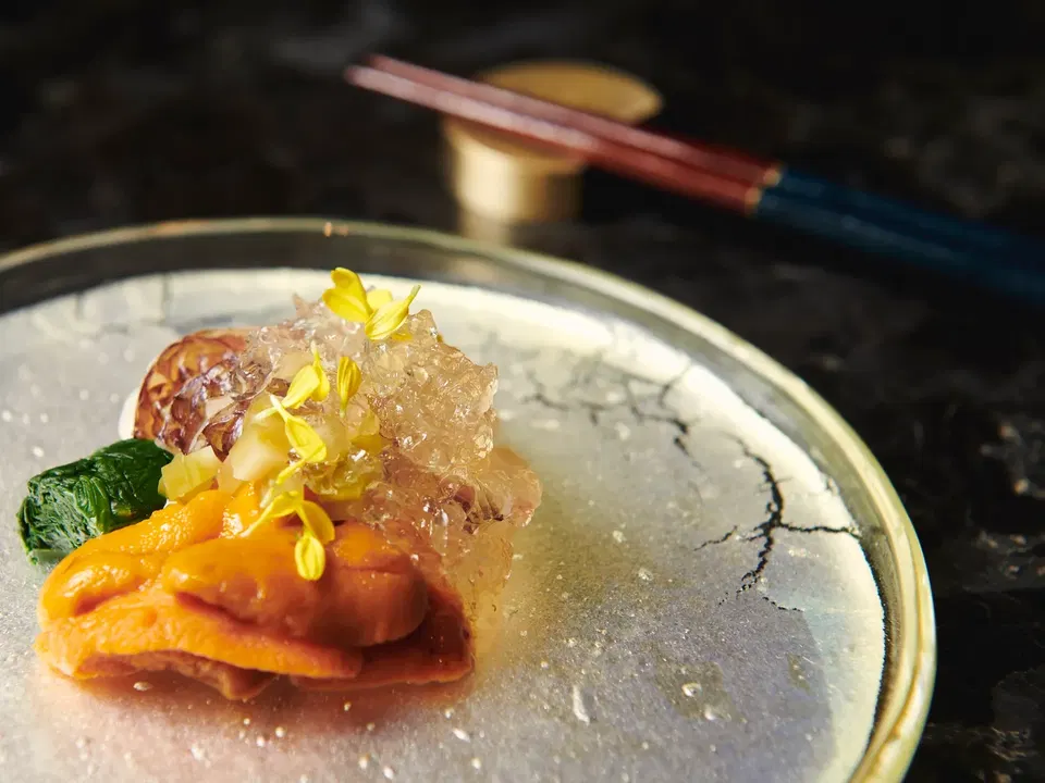A plated dish featuring layers of orange citrus segments, translucent ice or gelatin crystals, dark green leaf, octopus or seafood, and yellow flower